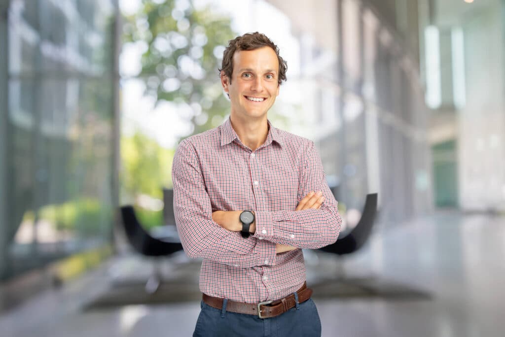 Professional corporate headshot of a smiling man in a checkered shirt, blue pants, and a brown belt, standing with arms crossed in a modern office setting featuring large glass windows and a blurred background of green foliage and sleek furniture. Captured by Linger Photography, showcasing a polished and approachable corporate image.