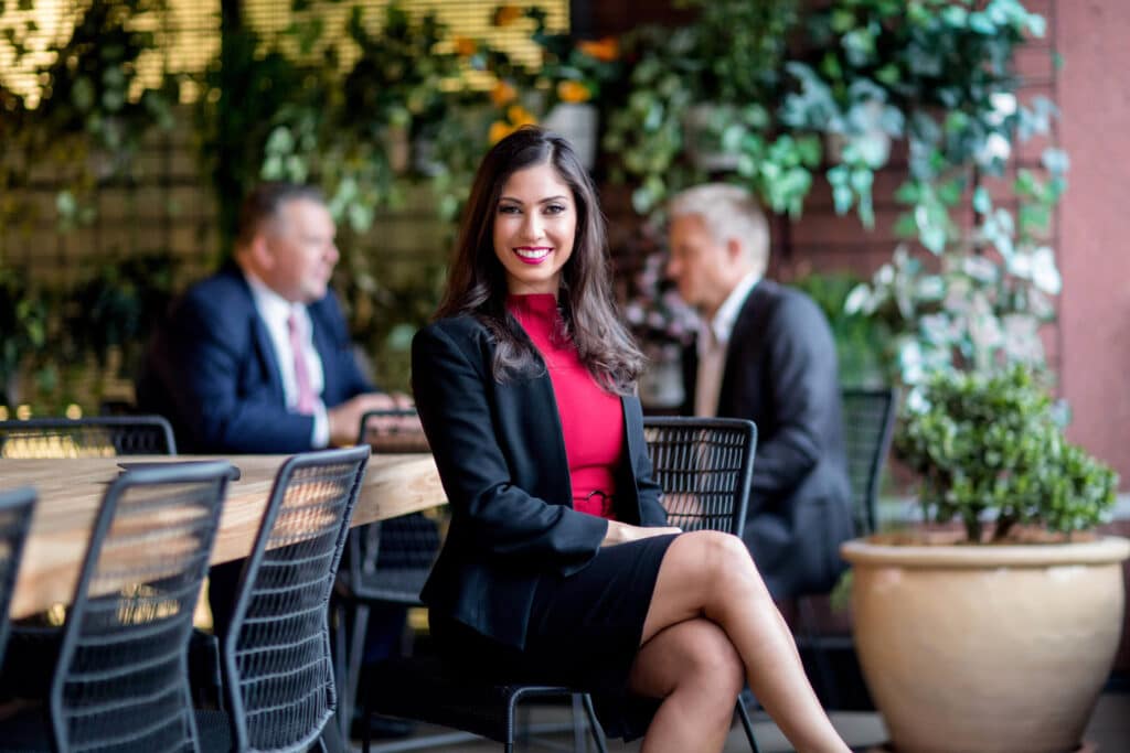 Professional corporate portrait of a confident woman in a black blazer and red top seated in an outdoor workplace setting with blurred greenery and two business professionals in the background. Captured by Linger Photography, emphasizing a polished and approachable professional image in a vibrant and dynamic environment.