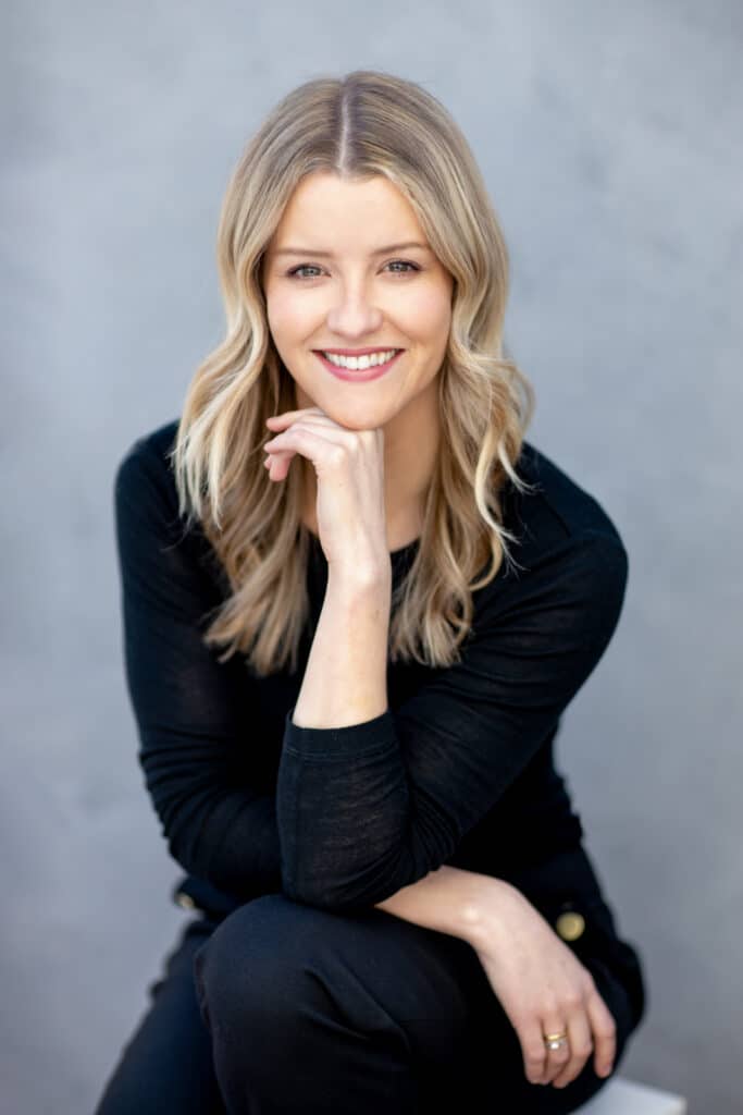 Professional individual headshot of a smiling woman with blonde wavy hair, wearing a black outfit, sitting with her chin resting on her hand. Captured outdoors against a soft, neutral background, exuding warmth, confidence, and approachability. A perfect blend of natural light and professional photography by Linger Photography.