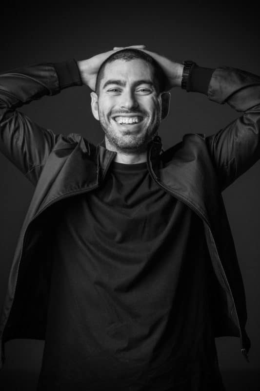Dynamic black-and-white individual headshot of a smiling man with hands resting behind his head, wearing a casual black jacket and t-shirt. Captured by Linger Photography, showcasing a relaxed and confident personality through modern studio lighting and minimalist design.