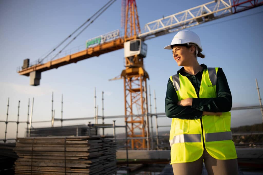 Professional business branding photograph of a female construction professional at a worksite captured by Linger Photography. She is wearing a high-visibility yellow vest and a white hard hat with 'Piety Group' branding, confidently standing with crossed arms against the backdrop of a construction crane and stacked building materials. The golden hour sunlight highlights her empowering stance and the dynamic construction environment.