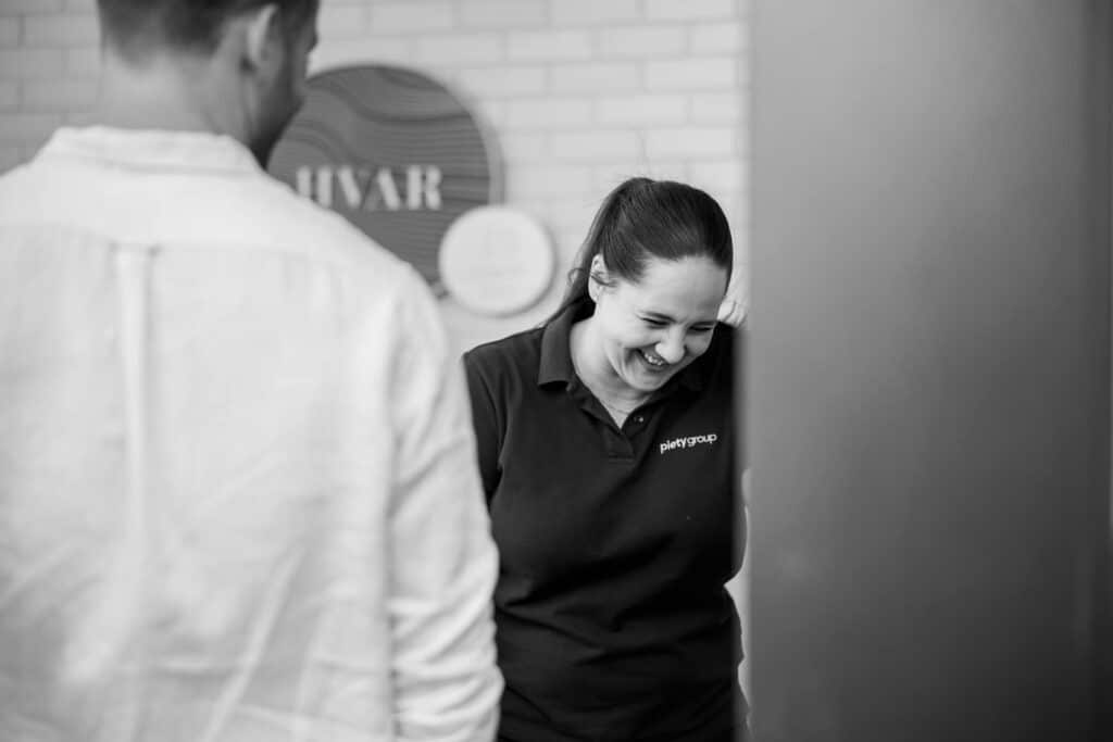 Black-and-white business branding photograph captured by Linger Photography, featuring a joyful moment of a professional woman wearing a branded polo shirt with 'Piety Group' embroidery. She is caught laughing naturally in a casual office environment, interacting with a colleague blurred in the foreground. The composition exudes authenticity, approachability, and team collaboration, ideal for showcasing company culture and values.