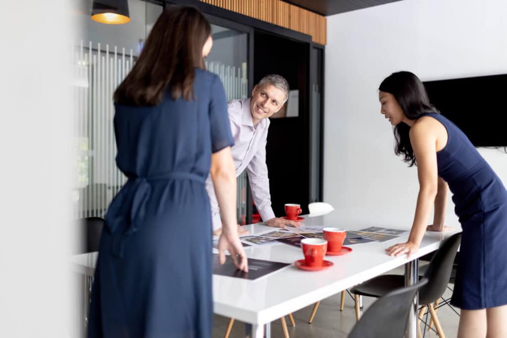 Office team collaboration with two women and one man engaged in discussion around a white table with red coffee cups, in a modern workspace featuring glass walls and wooden accents. Captured by Linger Photography, highlighting teamwork, creativity, and a professional atmosphere.