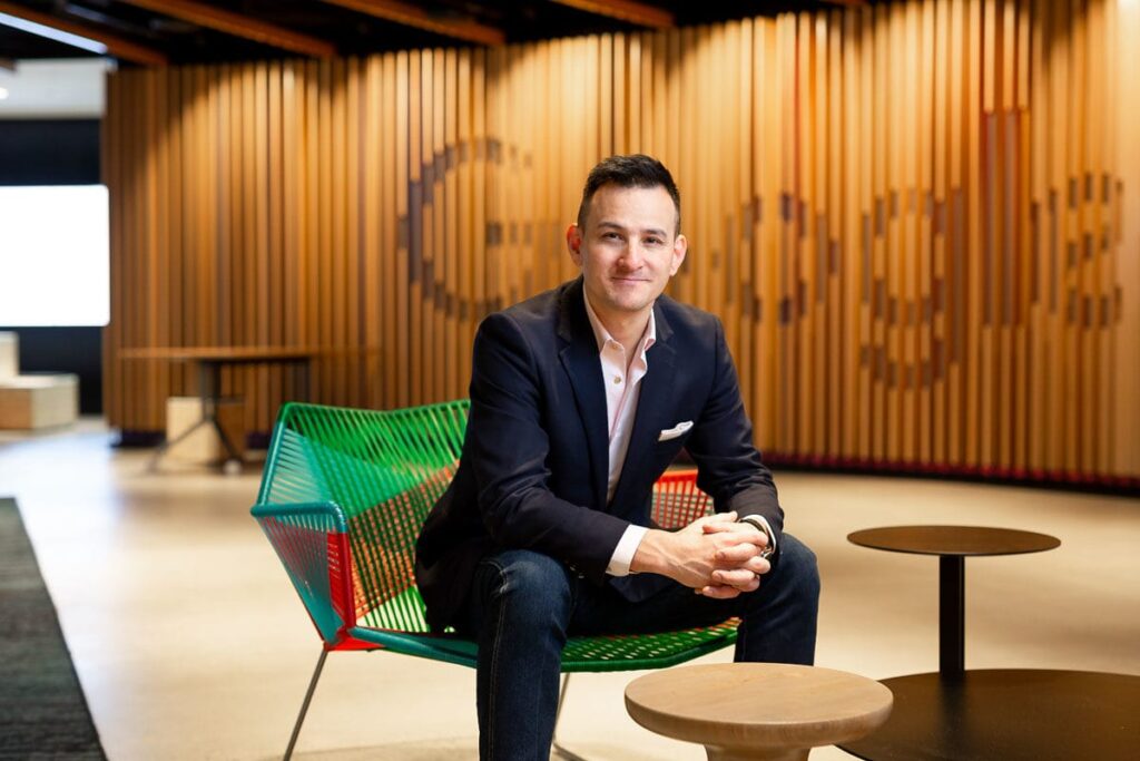 Professional portrait of a confident man seated on a modern green and red chair in a contemporary office space with a Google-branded wooden backdrop. Captured by Linger Photography, showcasing professionalism, creativity, and approachability.