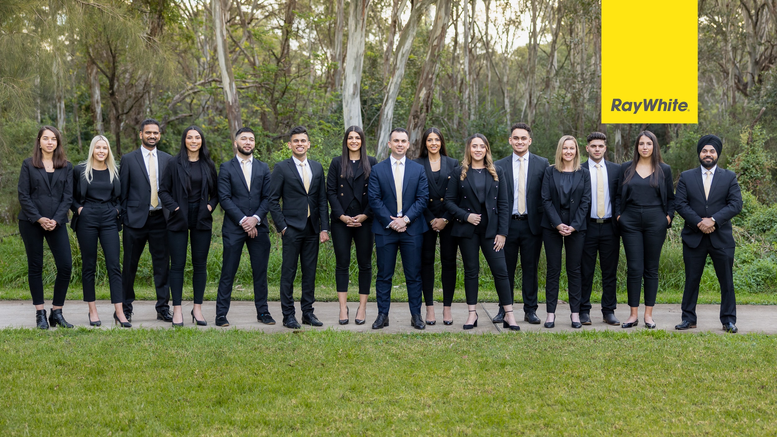 Group photo of the Ray White team standing outdoors in formal business attire with matching black suits and gold ties, set against a serene natural background of trees and greenery. Captured by Linger Photography, showcasing professionalism, unity, and corporate identity.